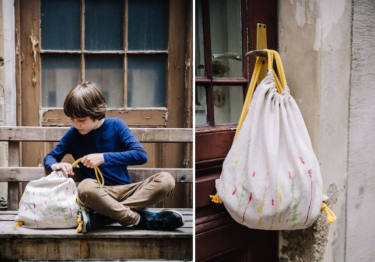 Junge sitzt mit seinem Turnsackerl auf einer Bank und zieht die Schnüre zu. Rechts im Bild hängt das Sackerl auf einer Türklinke außen am Haus.