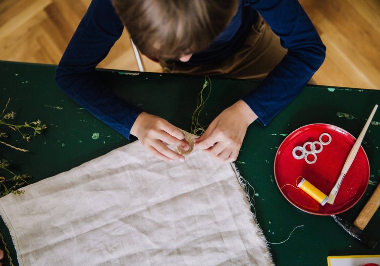 Bub sitz an einem grünen Tisch und näht mit Nadel und Faden die Seiten des Turnsackerls zusammen.