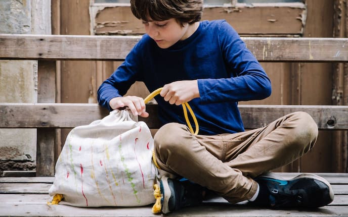 Bub sitzt auf einer alten Holzbank und zieht die Bänder seines selbstgemachten Turnsackerls zusammen.