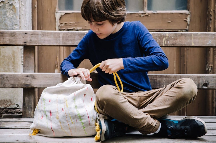 Bub sitzt auf einer alten Holzbank und zieht die Bänder seines selbstgemachten Turnsackerls zusammen.