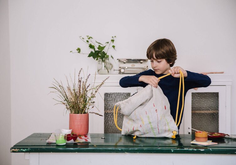 Der Junge fädelt die gelbe Schnur durch den Tunnel und die Ösen des Turnsackerls. Dabei steht der Turnsack auf der grünen Tischplatte vor einer weißen Wand vor der eine niedrige alte weiße Holzvitrine mit Glastüren steht.