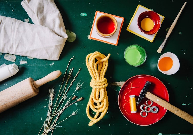 Auf einem grünen Tisch liegt ein Nudelwalker, Stoff fürs Turnsackerl, Butterpapier, gelbe Schnur, Farben, Pinsel, Nadel, Faden, Ösen und Stroh.