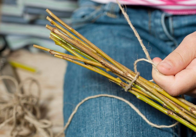 Nahaufnahme, eine Kinderhand, die um ein kleines Bündel Weidenzweige eine Schnur wickelt und sie Enden verknotet.