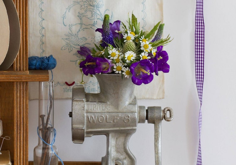 Fleischwolf umfunktioniert zu einer Blumenvase. Er ist an ein Regal geschraubt und in der Öffnung steck ein frischer Blumenstrauß aus weißen und violetten Blümchen.