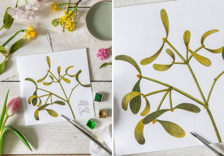 Tulpen, Narzissen liegen auf einem weißen rustikalen Holzboden, daneben liegt eine selbstgemachte Grußkarte mit einer gemalten Mistel und den Worten: Alles Gute. Pinsel und Farben liegen daneben.