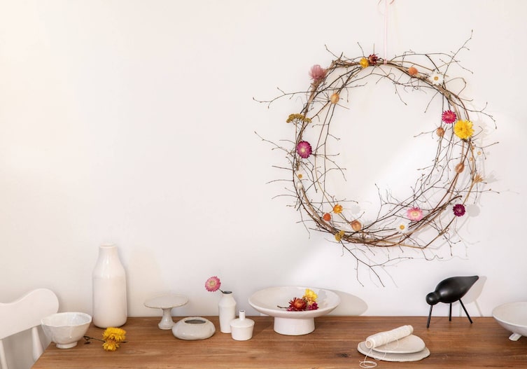 An einer weißen Wand hängt ein Kranz aus wenigen Zweigen mit bunten Trockenblumen. Darunter steht ein Holztisch mit weißen Keramiken.