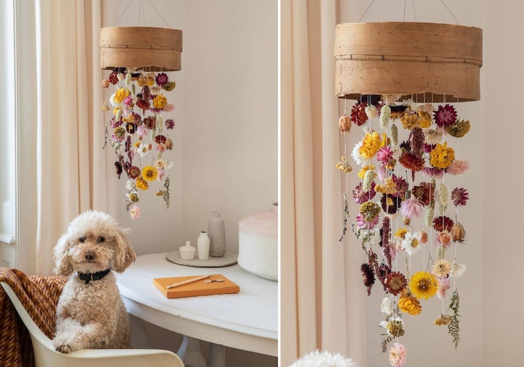Wohnecke in weiß von der Decke hängt ein altes Mehlsieb an dem Trockenblumen angebracht sind. Ein Tisch steht daneben mit einem Sessel auf dem ein weißer Pudel sitzt.