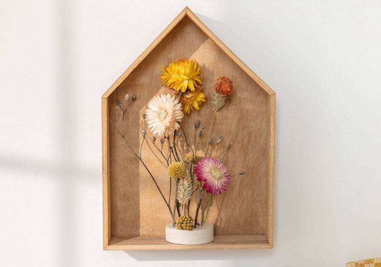 Ein kleines Holzhäuschen in dem bunte Trockenblumen stecken und gemeinsam an die Wand gehängt sind.