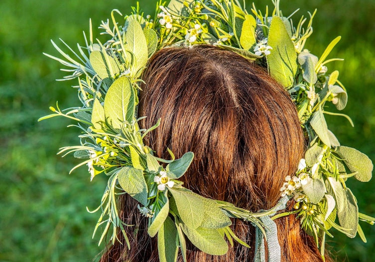 Frauenkopf mit dunklen langen Haaren von hinten. Sie hat einen Kräuterkranz auf mit weißen Blüten, Salbeiblättern und Lavendel.