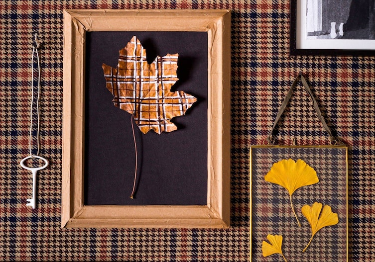 Braun karierte Tapetenwand, darauf hängt ein alter Schlüssel auf einem Band, ein Glasrahmen mit gepressten gelben Blumen und ein alter Rahmen, der mit Paketband aus Papier beklebt ist. Im Rahmen auf einem schwarzen Fotokarton ist ein Ahornblatt befestigt, das auch kariert, mit weißer und schwarzer Farbe bemalt ist.