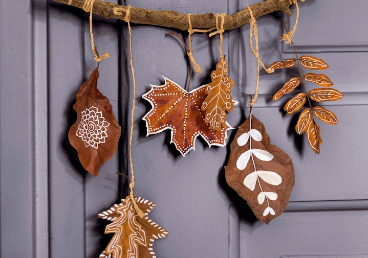 Graue Holztür, daran hängt ein dickerer Zweig an dem unterschiedlich getrocknete Herbstblätter mit verschiedenen Mustern, die mit weißem Kreidestift bemalt wurden, angebracht wurden.