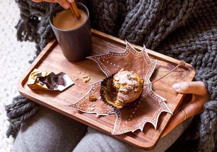 Frau sitzt gemütlich auf dem Boden in eine dicke graue Decke gewickelt, auf ihrem Schoß steht ein kleine Holzservierbrett, darauf liegt ein Stück eingewickelte dunkle Schokolade, ein Häferl mit Kakao, in den sie eine Stange Zimt taucht und ein bemaltes Ahornblautt auf dem ein Muffin steht.