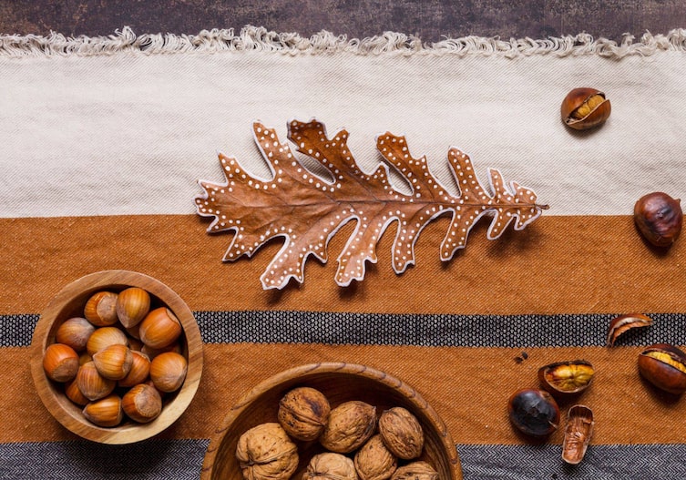 Tisch auf dem ein grau-weiß-braun gestreiftes Tischtuch liegt, darauf steht eine kleine Holzschüssel mit Haselnüssen in der Schale, eine etwas größere Holzschüssel mit Walnüssen in der Schale, lose liegen ein paar gebratene Maroni herum und mittig davon liegt ein ein großes Eichenblatt, das mit weißer Kreidefarbe bemalt ist. Die Ränder sind weiß umrandet und in der zweiten Reihe eine gepunktete Linie, ganz rundherum ums Blatt.