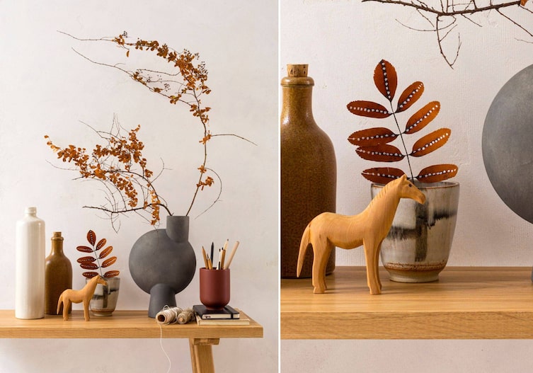 Ein Beistelltisch aus Holz. Darauf stehen unterscheidliche Keramikgefäße, Flaschen, und Vasen, ein Stiftehalter, zwei Notizbücher, ein kleine Holzpferd, in einer dunkelgrauen Betonvase steckt ein Zweig getrockneter Zweig mit vielen kleinen Blättern darauf. Daneben steckt in einem grauen Keramikhäferl ein getrockentes Rubinienblatt mit weißen Kreidepunkten