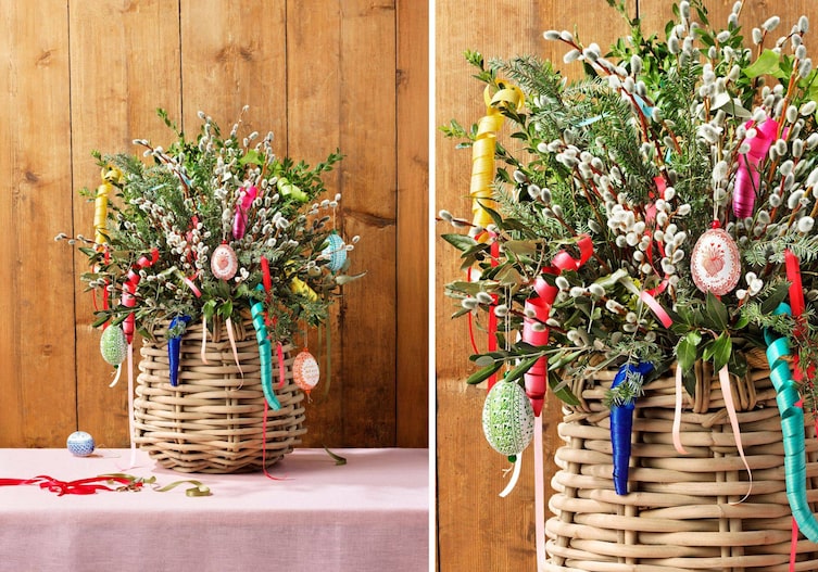 Tisch mit rosa Tischdecke vor einer Holzwand. Darauf steht ein Weidekorb, der mit Palmzweigen, Lorbeerzweigen und Olivenzweigen gefüllt ist. Darauf. hängen bunte Holzlocken und filigran bemalte Ostereier.
