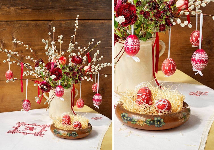 Eine weiße Weihkorbdecke liegt auf dem Tisch, daneben stent eine bemalte alte Holzschüssel mit Holzwolle und roten Ostereiern mit weißem Muster aus Wachs. Die Eier hängen auch am Osterstrauch mit Palmkätzchen und roten Frühlingsschnittblumen, Ranunkeln und Tulpen.