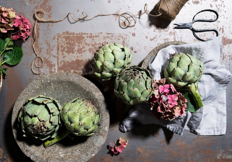 Alte Rostplatte von oben fotografiert, auf der Platte steht eine Steinschale in der zwei Artischocken liegen, daneben liegen weitere Artischocken und rosa Hortensienblüten.