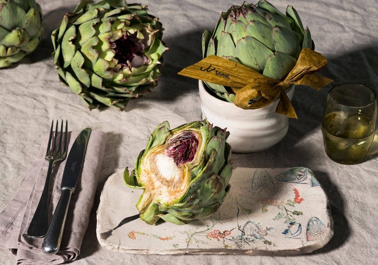 Gedeckter Tisch mit Naturfarben, Leinentischtuch, Leinenserviette, mehrere Artischocken, in einer Keramikschüssel steckt eine Artikschocke, drumherum ist ein Band gewickelt auf dem der Name der Tischkarte steht.