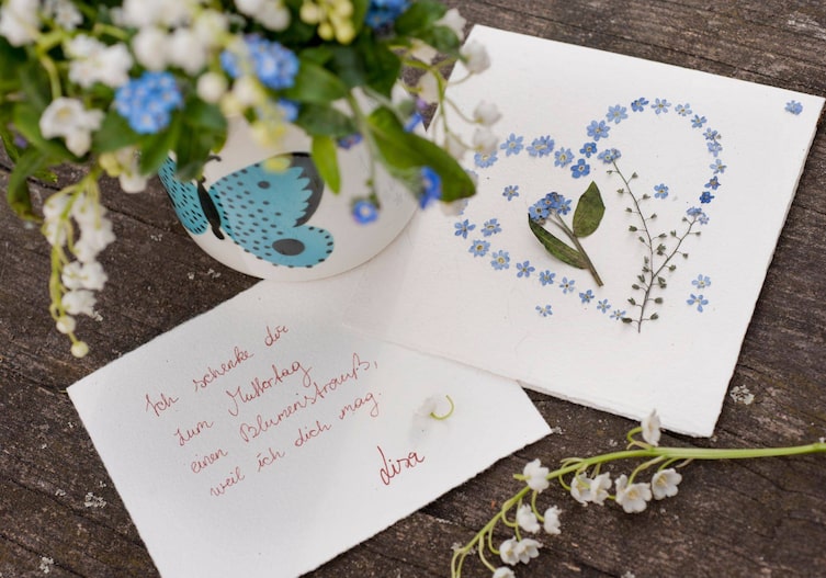 Selbst gebastelte Grußkarte, auf der Vorderseite ist aus getrockneten Vergissmeinnichtblüten ein Herz gelegt. Auf der anderen Grußkarte steht: Ich schenke dir zum Muttertag einen Blumenstrauss, weil ich dich mag. Lisa. Daneben steht ein weißes Tongefäß mit einem türkisen Schmetterling, darin stecken Maiglöckchen und Vergissmeinnicht.