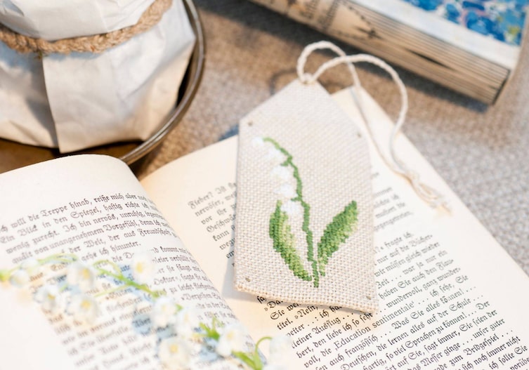 Altes offenes Buch auf dem ein Leseszeichen aus Naturleinen liegt, das mit einem Maiglöckchen bestickt ist.