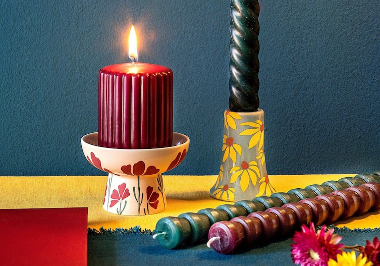 Weißer Kerzenhalter mit rotem Blumenmuster und dunkelroter gerillter, brennenden Stumpenkerze. Daneben steht ein blau-beiger Kerzenstender mit gelben Blumenmotiv und einer gedrehten grünen Stabkerze.