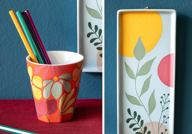 Keramiktrinkbecher orange bemalt mit aufgemalten Blumenmotiven. Er dient als Stiftehalter. An der Wand hängt ein weißes Keramiktablett mit Zweigen und bunten großen Punkten bemalt.