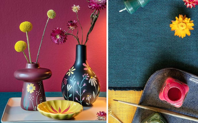 Blumenvasen, weinrot und schwarz, mit Blumenmotiven bemalt. Gelbe Keramikschüssel mit roter Blume. Daneben sieht man Keramikfarben und einen Pinsel auf einem Keramiktablett. Getrocknete Blumen und einen Teil einer grünen Stabkerze