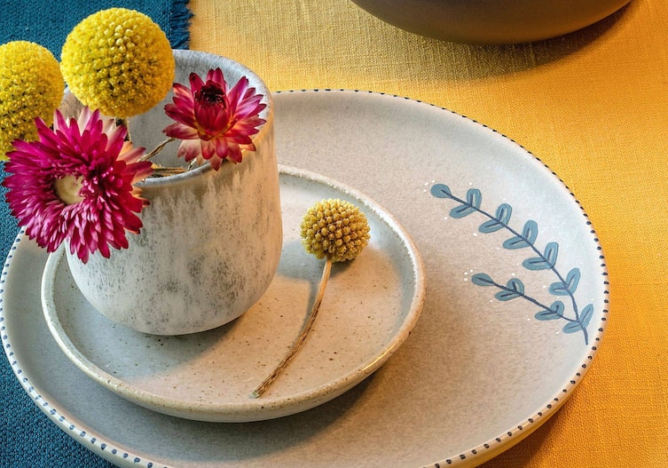 großer Keramikteller, der am Rand mit einem blauen Zweig bemalt ist. Darauf steht ein kleiner Untersetzer und ein Keramikbecher im selben Stil. Der Becher ist mit gelben und dunkelrosa Blumen geschmückt.