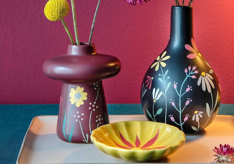 Zwei bauchige Vasen, eine weinrot, die andere schwarz, mit Keramikfarbe bunte Blumen aufgemalt. Davor steht eine kleine gelbe mit einer roten Blume bemalten Keramikschüssel. Alle drei Objekte stehen auf einerm weißen Keramikobjekt