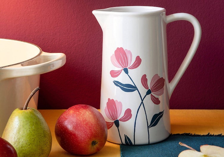 Weißer Wasserkrug aus Keramik, mit drei rosa Blüten und Stängeln bemalt. Daneben liegt ein roter Apfel und eine grüne Birne.