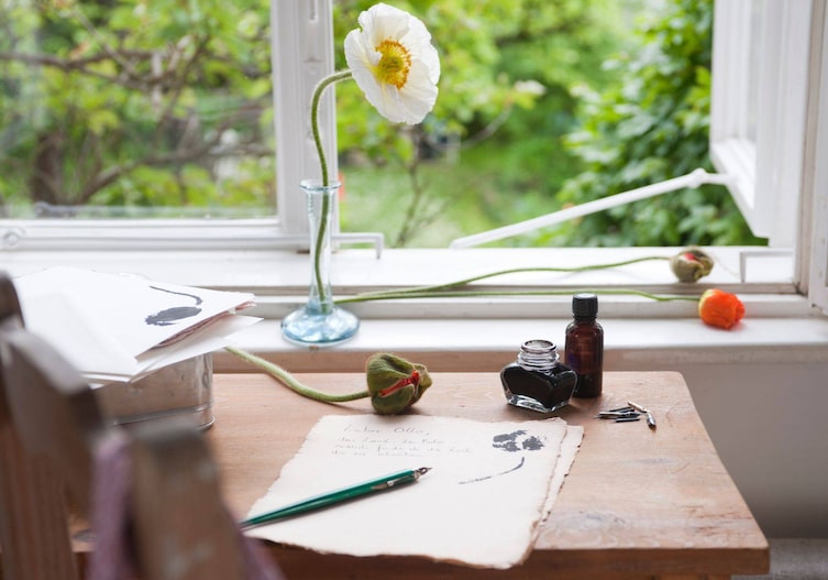Ein uriger Holzschreibtisch, der am Fenster steht. Das Fenster ist offen, draußen blüht alles und ist grön. Auf dem Fensterbrett steht eine kleine Glasvase mit einer weißen Monblume darin. Auf dem Tisch liegt altes Papier mit einer grünen Schreibfeder, ein Tintenfass mit unterschiedlichen Schreibspitzen für die Feder liegt daneben. Auf dem Briefpapier ist ein Abdruck mit Tinte von einer Monblumenknospe zu sehen. Neben dem Blatt liegt auch eine Mohnblumenknospe.