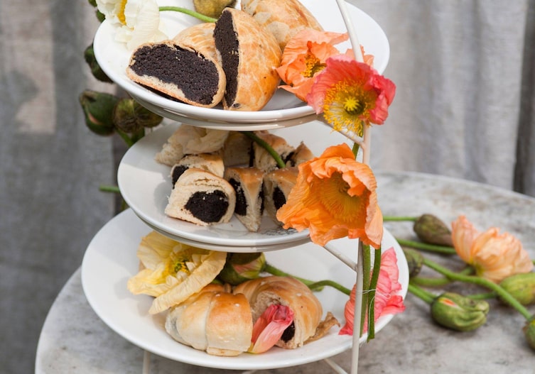 Etagere mit weißen Keramiktellern, darauf liegen aufgeschnittene Mohnzelte, die mit frischen roten, orangen, gelben und weißen Mohnblumen geschmückt sind.