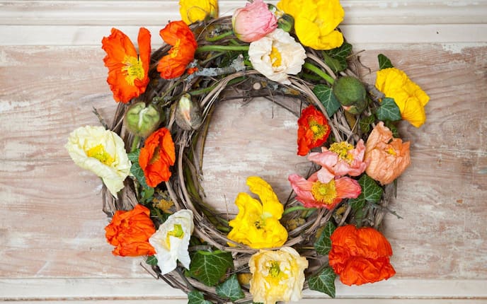 Türkranz aus Zweigen, mit gelben, roten und weißen Mohnblumen geschmückt und mit Efeu verziert. Der Kranz liegt auf einem abgebeizten weißlichen Holzuntergrund.