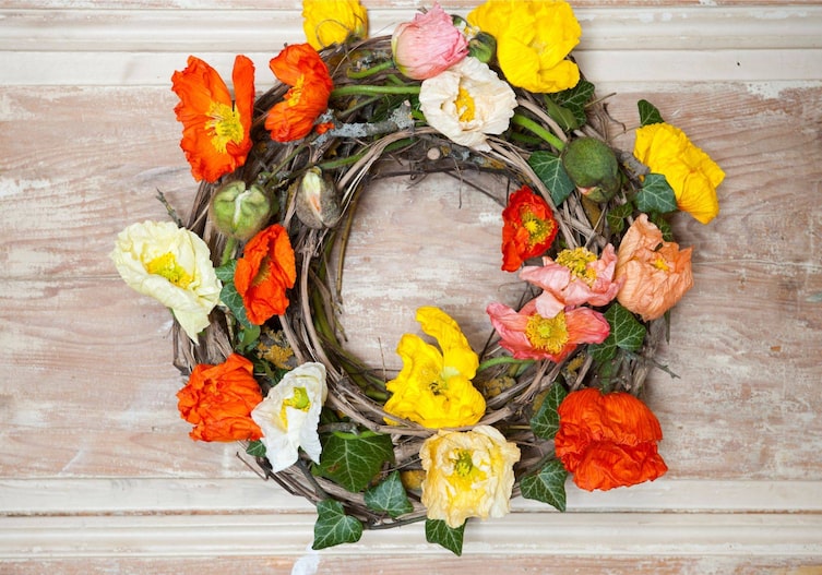 Türkranz aus Zweigen, mit gelben, roten und weißen Mohnblumen geschmückt und mit Efeu verziert. Der Kranz liegt auf einem abgebeizten weißlichen Holzuntergrund.
