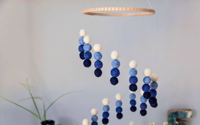 DIY-Mobile aus Filzkugeln in Blau- und Cremetönen, aufgehängt an einem Holzreifen, mit Bastelutensilien auf einem weißen Tisch darunter.