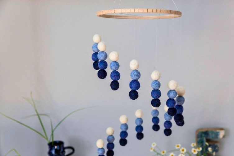 DIY-Mobile aus Filzkugeln in Blau- und Cremetönen, aufgehängt an einem Holzreifen, mit Bastelutensilien auf einem weißen Tisch darunter.