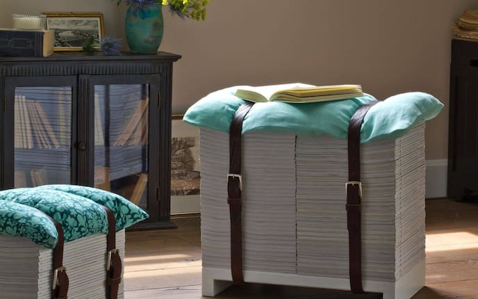 Wohnzimmer, auf dem braunen Holzboden stehen zwei Hocker aus Servus Magazinen, die Hefte stehen auf einem weißen Holzkonstrukt und sind mit zwei alten Ledergürteln gebunden. Als Sitzfläche dient ein türkiser Polster.