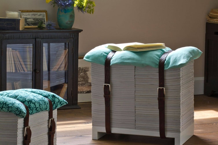 Wohnzimmer, auf dem braunen Holzboden stehen zwei Hocker aus Servus Magazinen, die Hefte stehen auf einem weißen Holzkonstrukt und sind mit zwei alten Ledergürteln gebunden. Als Sitzfläche dient ein türkiser Polster.