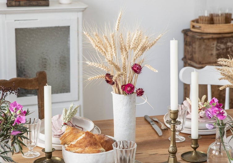 In Naturfarben gedeckter Tisch mit Messingkerzenständern und einer Vase in der Ähren und Strohblumen stecken.