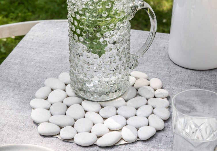 Untersetzer mit weißen Steinen beklebt, darauf steht ein Glaskrug mit Wasser und Kräutern, alles steht auf einem Tisch in einem Garten.