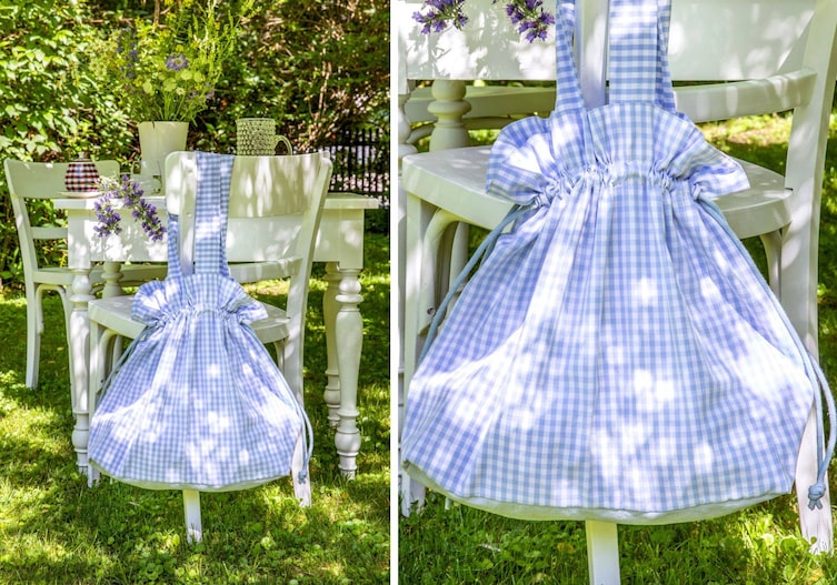 Weißer alter Tisch mit alten weißen Sesseln stehen in einem Garten in der Wiese. Es ist sommer. Über einer Sessellehne hängt eine große blau-weiß karierte Beuteltasche mit blauen Kordeln