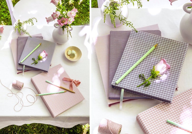 Ein Tisch mit weißer Tischdecke steht in einem Garten. Es ist Sommer. Auf dem Tisch liegen unterschiedliche Bücher, die von rosa bis violetten Karomuster-Stoffen beklebt wurden. Es liegen noch Bleistifter herum und eine rosa Schnur.