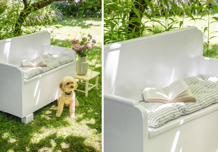 Grau-weiß karierte Sitzauflage mit einem aufgeschlagenem Buch auf einer weißen Holzbank, die im Schatten eines Baumes im Garten steht. Es ist Sommer, alles blüht. Vor der Bank sitzt ein weißer Pudel. Neben der Bank steht ein altes weißes Stocker, auf dem eine graue Vase mit Pfingstrosen und anderen Sommerblühern steht.