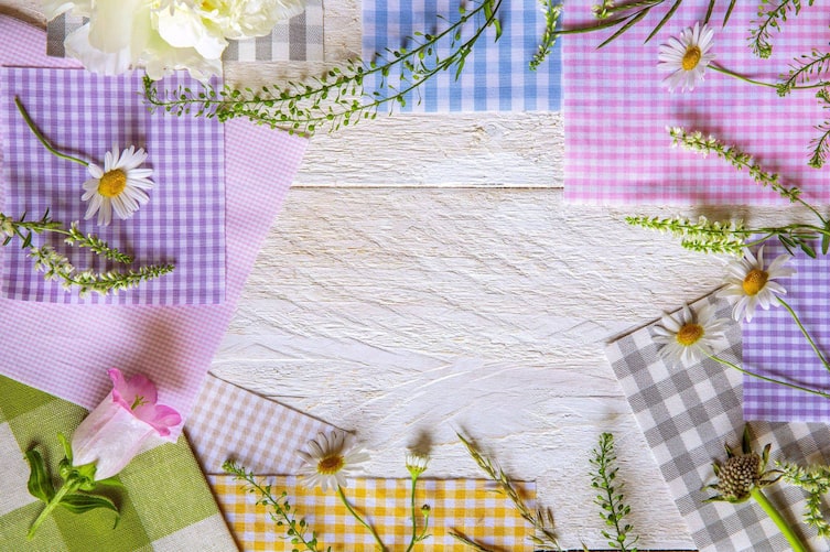 Bunt karierte Stoffreste liegen auf einem weiß gestrichenem Holzuntergrund. Auf den Stoffresten liegen noch verschiedene Sommerblumen.