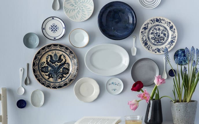 weiße und blaue alte, große und kleine Keramikteller und Keramiklöffel an der Wand, weißer Tisch, Bllumenvase, Blumentopf mit blauer Hyazinthe, Buch, Glas mit Saft