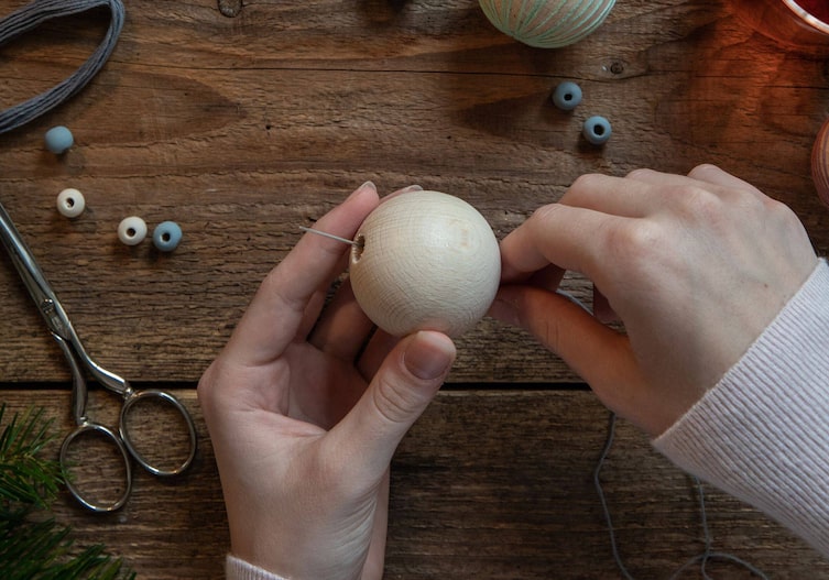 Zwei Frauenhände führen eine lange Nähnadel mit Stickgarn durch die Öffnung der Holzkugel
