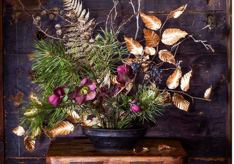 Föhrenzweige, Schneerose, goldbesprühter Farn und Blätterzweig sind in einem Gesteck in einem Topf gebunden, das auf einem kleinen Holzstockerl vor einer dunklen Holzwand steht.