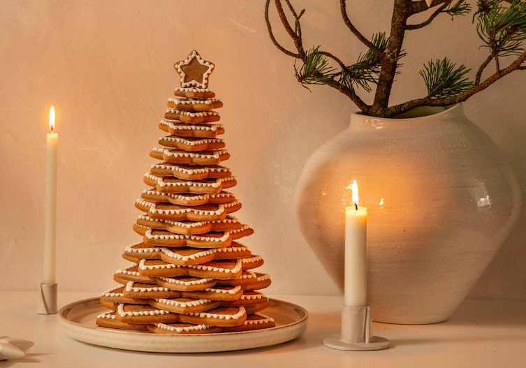 In unterschiedlichen Größen übereinanergestapelte Lebkuchensterne mit Zuckergussverzierung. Sie stellen einen Christbaum dar. Der Chrsitbaum steht auf einem weißen Keramikteller neben dem zwei weiße Stabkerzen brennen und eine weiße Keramikvase steht, in der ein Tannenzweig steckt.