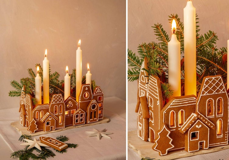Ein kleines Dorf aus verzierten Lebkuchenplatten. Sie sind im Viereck aufgebaut und darin stehen unterschiedlich lange, brennende Stabkerzen, die noch mit Tannenzweigen geschmückt sind.