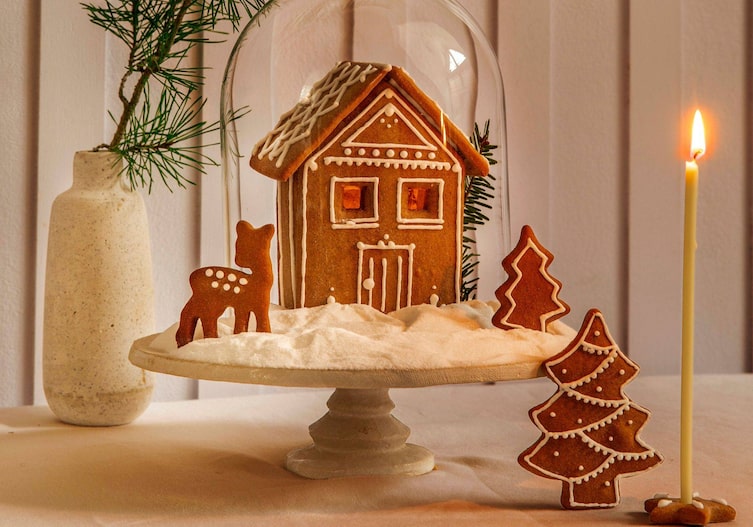 Ein selbstgebasteltes Lebkuchenhaus, mit Zuckerguss verziert steht unter einer Glaskuppel und steht auf einem Tortenteller das mit Zucker (als Schnee) verziert ist, auf dem ein Reh und ein Baum aus Lebkuchen steht.